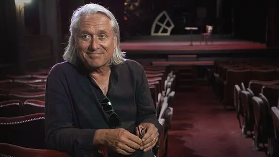 Der Regisseur Wilfried Hauke sitzt mit dem Rücken zur Theaterbühne und lächelt. | Wilfried Hauke Der Regisseur Wilfried Hauke sitzt mit dem Rücken zur Theaterbühne und lächelt.