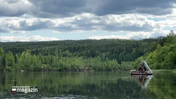 Ein Holzfloß mit Zelt drauf schwimmt über eine Wasseroberfläche.