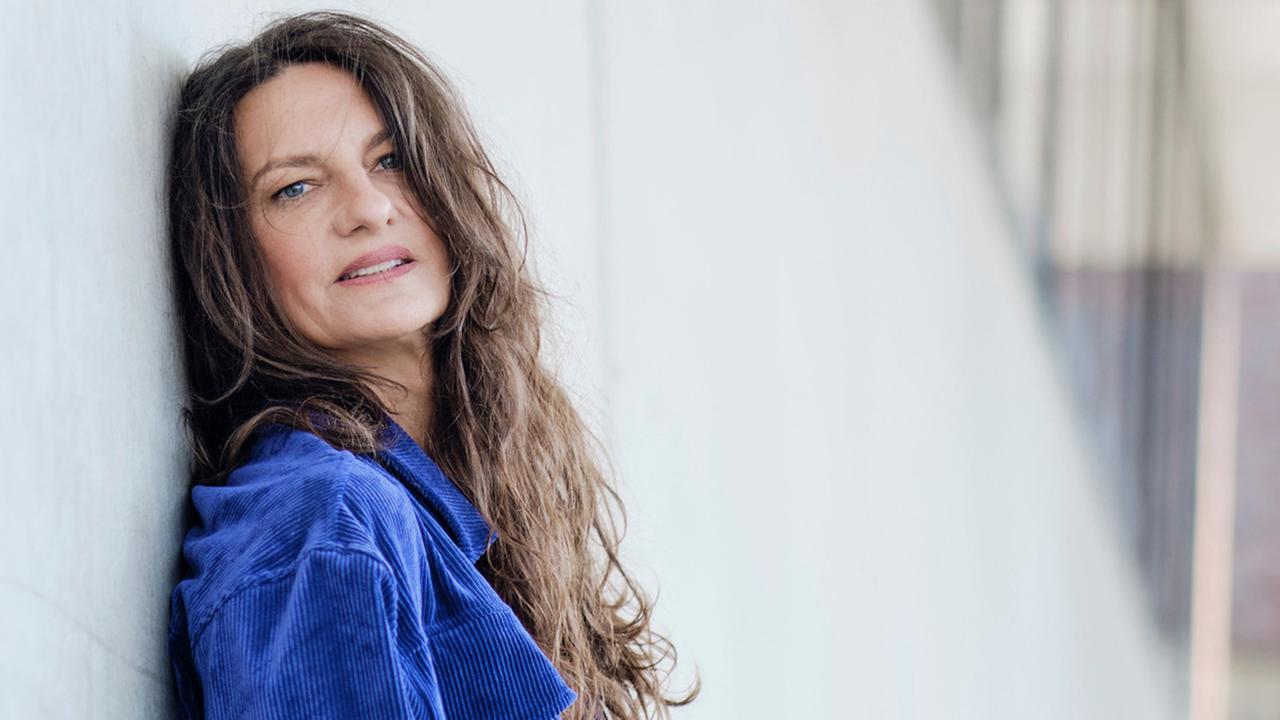 Catrin Striebeck lehnt an einer weißen Wand und posiert für die Kamera. 