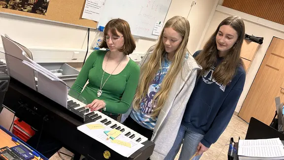 Drei Mädchen stehen an einem E-Piano. Die eine sitzt daran und drückt einige Tasten runter.