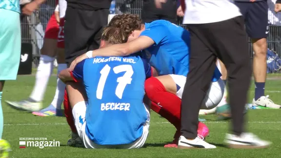 Nach einem verlorenen Spiel sitzt ein Spieler des Vereins Holstein Kiel mit der Nummer 17 und dem Namen "Becker" auf dem Rasen und wird von einem Mitspieler getröstet.
