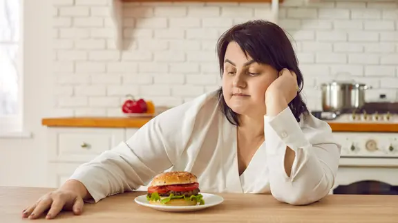 Binge-Eating-Störung: Frau sitzt am Tisch und starrt einen Burger an.