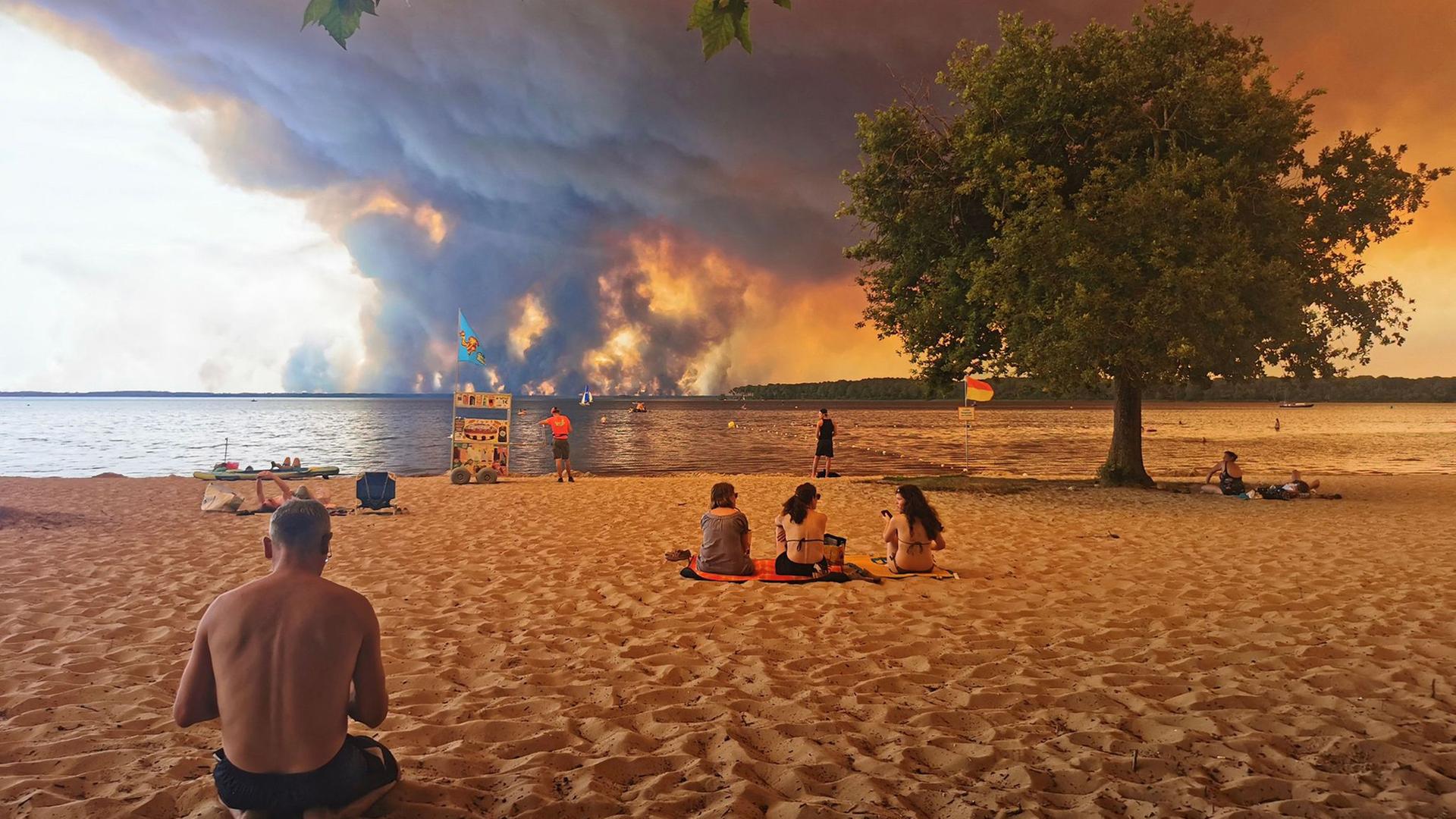 Strandbesucher sitzen am Strand von Moutchic in Lacanau im Südwesten Frankreichs, während Rauchwolken von einem Waldbrand in den Himmel aufsteigen. | AFP/dpa, Maximilien Lamy