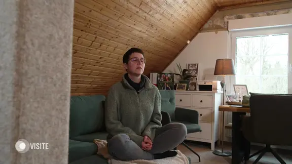 Eine Person sitzt im Schneidersitz auf einem grünen Sofa in einem gemütlichen Dachzimmer mit Holzdecke.