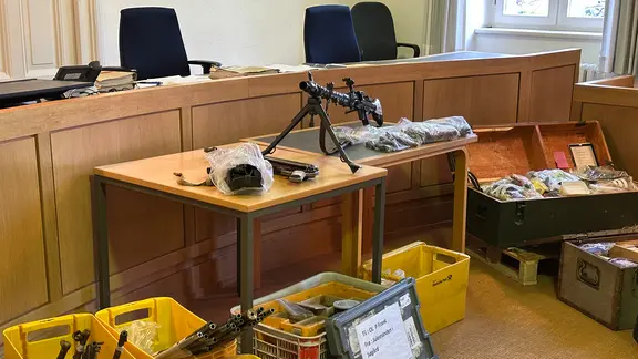 Eine Schusswaffe und Kisten mit weiteren Beweismitteln liegen in einem Gerichtssaal. | NDR, Ben Armstrong Eine Schusswaffe und Kisten mit weiteren Beweismitteln liegen in einem Gerichtssaal.