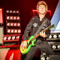 Billie Joe Armstrong von Green Day steht während eines Konzerts auf der Bühne des Hurricane Festivals. Er spielt eine grellgrüne E-Gitarre, trägt eine schwarze Hose und eine dunkle Jacke, und ist mitten in einer dynamischen Pose. Im Hintergrund sind leuchtende Bühnenlichter und Lautsprecher zu sehen.
