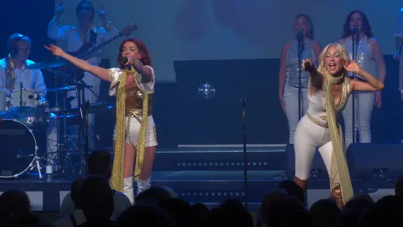 Zwei Frauen auf einer Bühne in Siebziger-Jahre-Kostümen animieren ein Publikum