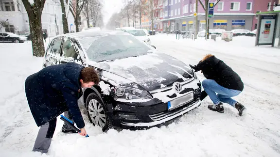Zwei Personen entfernen Schnee vor den Rädern eines festgefahrenen Auto.