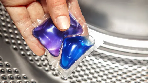 Ein Gel-Cup mit einer farbigen Waschflüssigkeit wird vor einer Waschtrommel gehalten. | Colourbox, ssuaphoto Ein Gel-Cup mit einer farbigen Waschflüssigkeit wird vor einer Waschtrommel gehalten.