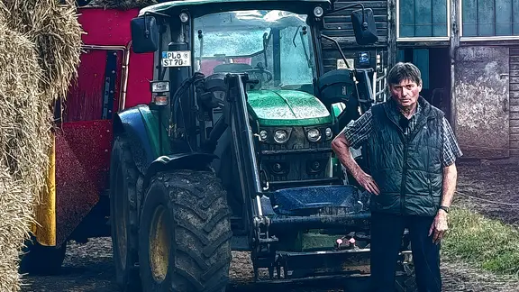 Landwirt aus der Doku vor einem großen Traktor. Scheune im Hintergrund. 