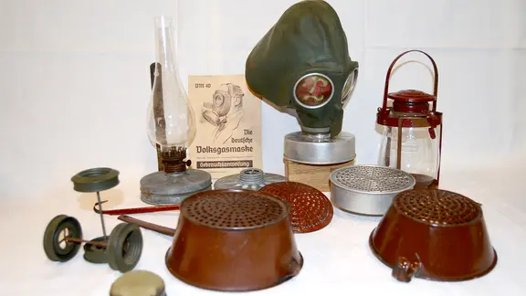 Auf dem Bild sind mehrere Gegenstände abgebildet, die aus den Filtergehäusen von "Volksgasmasken" gefertigt wurden: Petroleumlampen, Siebe, Schaumlöffel. Auch eine Gasmaske ist zu sehen.