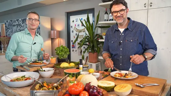 Fernsehkoch Tarik Rose (rechts) und Ernährungsmediziner Dr. Matthias Riedl stehen in einer Küche und essen Antipasti.