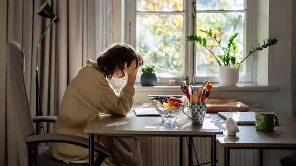 Eine erschöpfte Frau sitzt vor einem Fenster an einem Tisch und stützt den Kopf in die Hände.