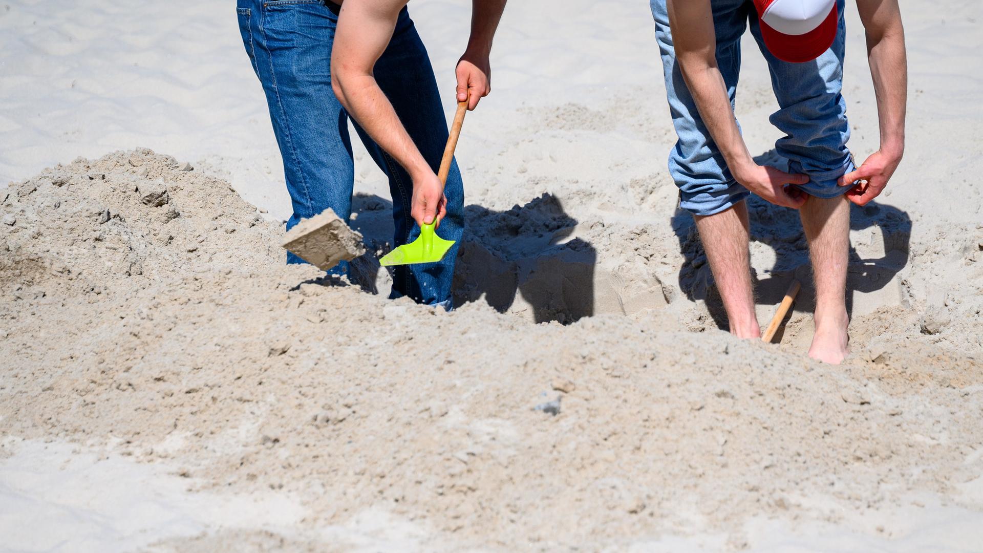 Zwei Strandbesucher buddeln im Sand, einer hält eine Schaufel. | picture alliance/dpa | Jonas Walzberg