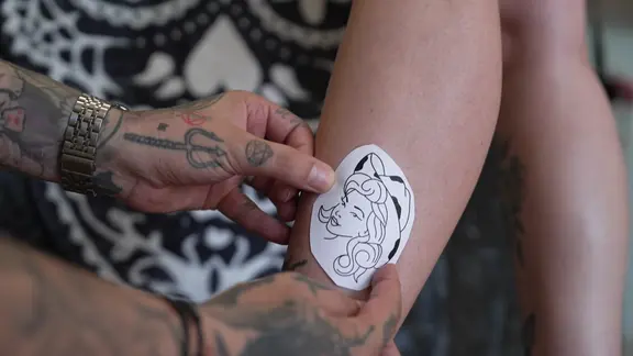 Zwei Hände halten eine Tattoovorlage auf der ein Frauengesicht mit Cowboy-Hut zu sehen ist auf ein Bein.