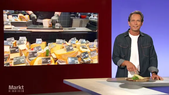 Moderator Jo Hiller im Studio von Markt, er zeigt auf ein Stück Käse, das vor ihm auf einem Tresen steht.