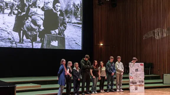 Eine kleine Gruppe von Schülerinnen und Schülern steht auf einer Bühne. Sie präsentieren Ergebnisse eines Workshops. Im Hintergrund sieht man auf der Leinwand ein Foto in schwarz-weiß mit Frauen auf der Flucht.