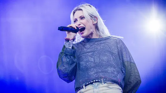 Emily Armstrong, die neue Sängerin von Linkin Park, singt leidenschaftlich auf der Bühne während des Konzerts in Hannover. Sie trägt ein glitzerndes, transparentes Oberteil und hält ein Mikrofon in der rechten Hand. Der Hintergrund ist in blaues Licht getaucht.