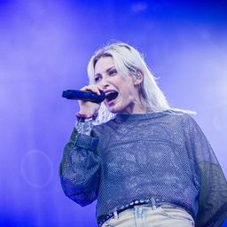 Emily Armstrong, die neue Sängerin von Linkin Park, singt leidenschaftlich auf der Bühne während des Konzerts in Hannover. Sie trägt ein glitzerndes, transparentes Oberteil und hält ein Mikrofon in der rechten Hand. Der Hintergrund ist in blaues Licht getaucht.