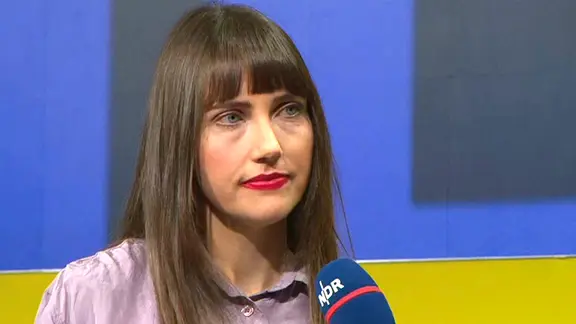 Μια γυναίκα με μακριά καστανά μαλλιά μιλάει στο μικρόφωνο