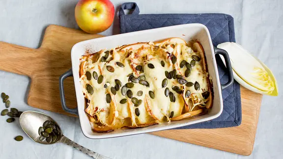 Auf einem Tisch liegt ein Holzbrett mit einem Topflappen und einer Auflaufform. Darin befindet sich ein schneller Chicorée-Apfel-Auflauf mit Kürbiskernen.