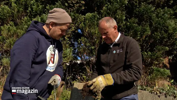 Samir Chawki und Thomas Balster begutachten eine Pflanze in einem Garten.