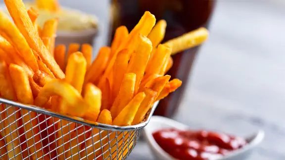 Vor dem Frittieren kann man die Pommes einige Minuten in kaltes Wasser legen. | Colourbox, Juan Moyano Ein Körbchen mit frisch zubereiteten Pommes frites, im Hintergrund ein Schälchen mit Ketchup.
