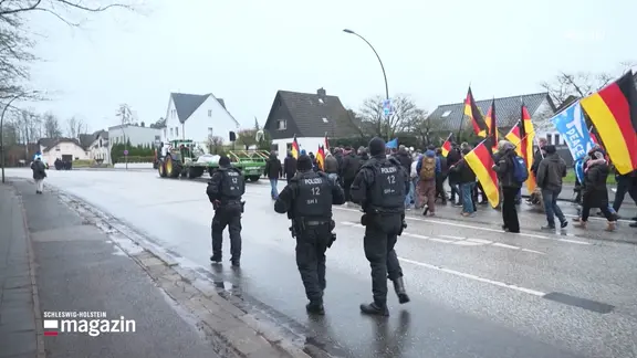 Ein Demonstrationszug von als rechtsextrem einzustufenden Personen, sie tragen Deutschlandflaggen. Neben dem Demonstrantenzug laufen Polizisten in Schutz-Montur.