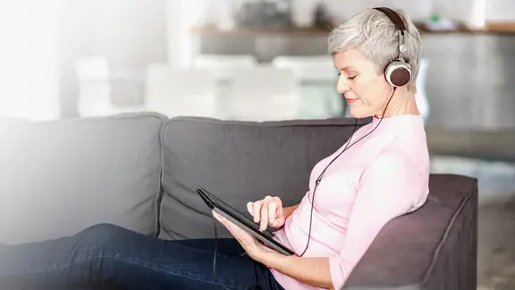 Frau mit Kopfhörern sitzt auf dem Sofa mit einem Laptop auf dem Schoß.