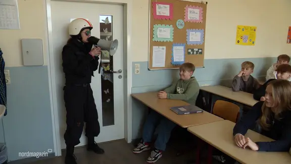 Eine verkleidete Person mit Helm und Megaphon steht in einem Klassenzimmer.