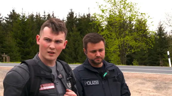 Ein Motorrad fahrer steht mit einem irritierten Blick neben einem Polizisten.