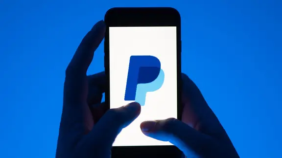 Eine Hand hält ein Smartphone mit dem Logo des Bezahldienstes Paypal. | Sebastian Kahnert/dpa Eine Hand hält ein Smartphone mit dem Logo des Bezahldienstes Paypal.