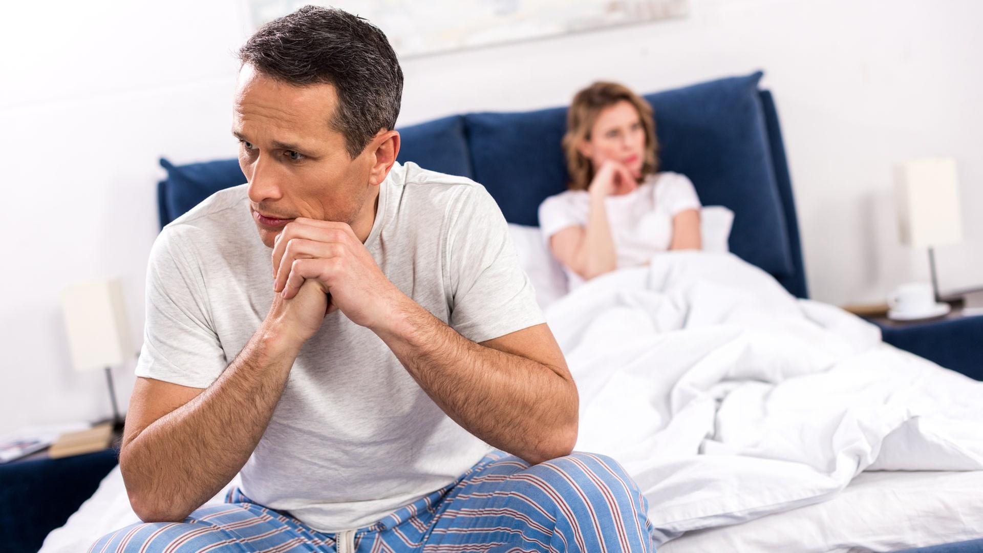 Ein Mann sitzt auf einem Bett, seine Frau ist unscharf im Hintergrund zu sehen. | Colourbox, -