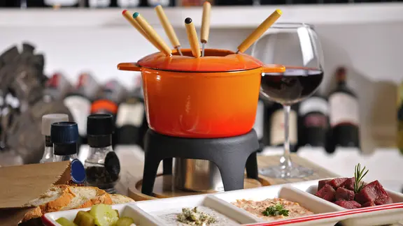 Ein orangefarbener Fonduetopf auf einem Rechaud, davor steht ein länglicher Teller mit Soßen, Fleisch und Gurken. | fotolia, COSPV Ein orangefarbener Fonduetopf auf einem Rechaud, davor steht ein länglicher Teller mit Soßen, Fleisch und Gurken.