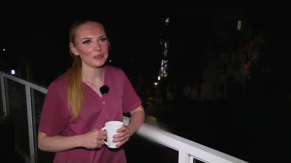 Eine junge Frau mit blonden Haaren und einem brombeerfarbenen Kittel hält eine Tasse in den Händen.