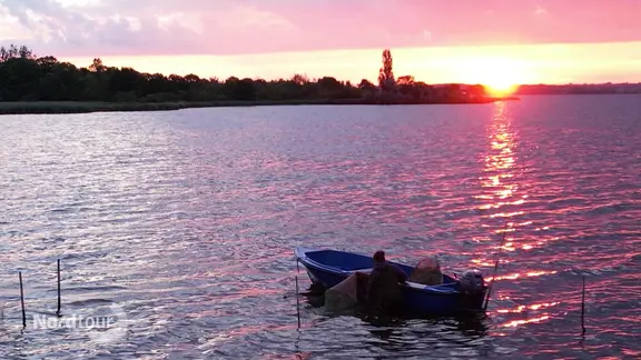 Bei Sonnenaufgang schwimmt ein kleines Beiboot in einer Bucht neben einem Krabbenfischer in Wathose.