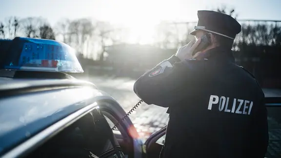 Ein Polizist in Uniform steht neben einem Polizeiauto und hält einen Telefonhörer in der Hand.