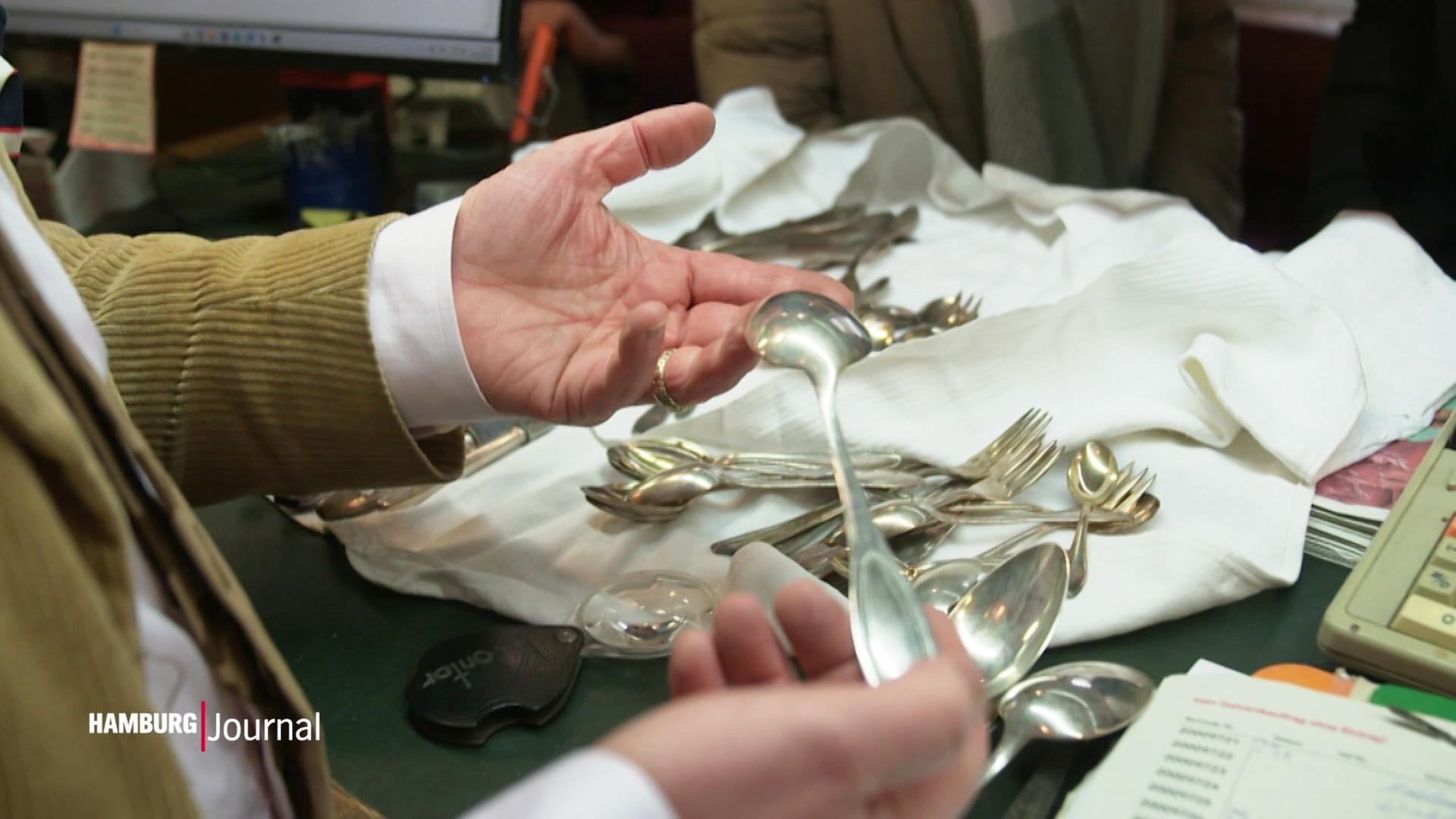 Zwei Hände halten einen Silberlöffel, auf einem Tisch liegt weiteres Besteck aus Silber. | Screenshot