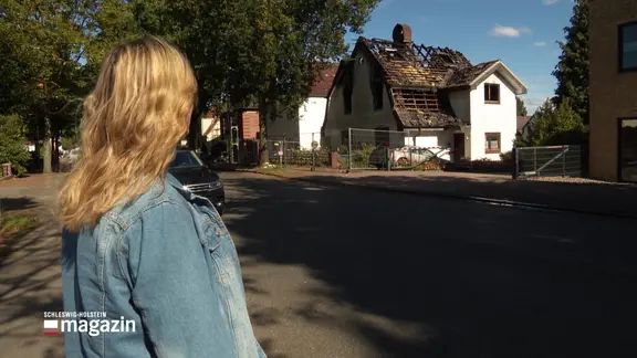 Eine Frau blickt von der gegenüberliegenden Straßenseite auf ein ausgebranntes Wohnhaus.