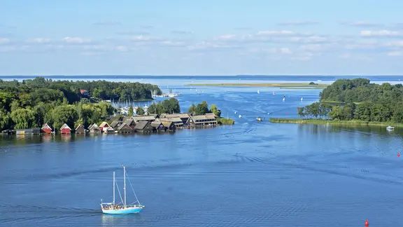 Ein Segelboot fährt bei Röbel auf der Müritz, am Ufer Bootshäuser.