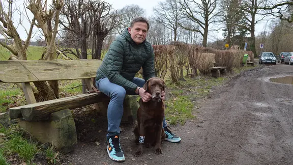 Ein Mann sitzt auf einer Bank neben seinem dementen Hund und blickt in die Kamera.