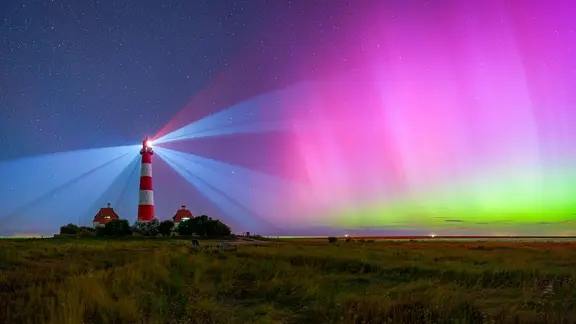 Ein rot-weiß gestreifter Leuchtturm, der in Betrieb ist, wird von violett grünen Polarlichtern angeschienen.