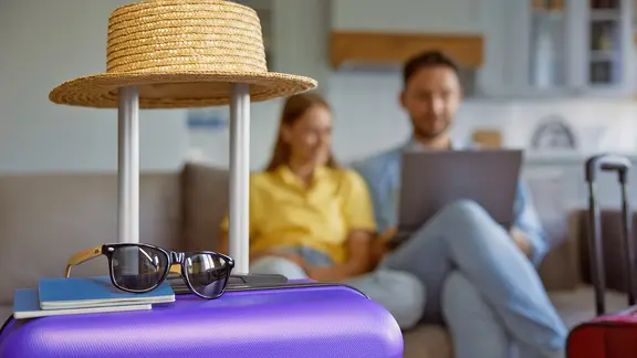 Koffer, Pässe, Sonnenhut und Sonnenbrille, im Hintergrund ein Paar mit Laptop