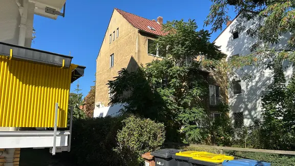 Vor einem dreistöckigen Wohnhaus stehen mehrere hohe Bäume.