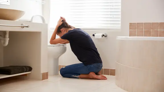 Erbrechen: Eine Frau kniet im Badezimmer vor der Toilette. Sie hält wie beim Erbrechen den Kopf über die Kloschüssel und bindet die langen Haare mit den Händen nach hinten.