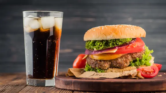Ein Glas Cola mit Eiswürfeln steht auf einem Tisch. Daneben ein Tablett, auf dem ein Hamburger angerichtet ist.
