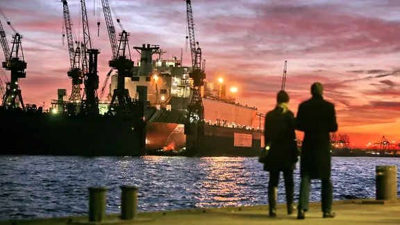 Ein Paar steht an den Landungsbrücken im Hamburger Hafen und blickt auf die Elbe mit dem Schwimmdock und den stimmungsvollen Abendhimmel.
