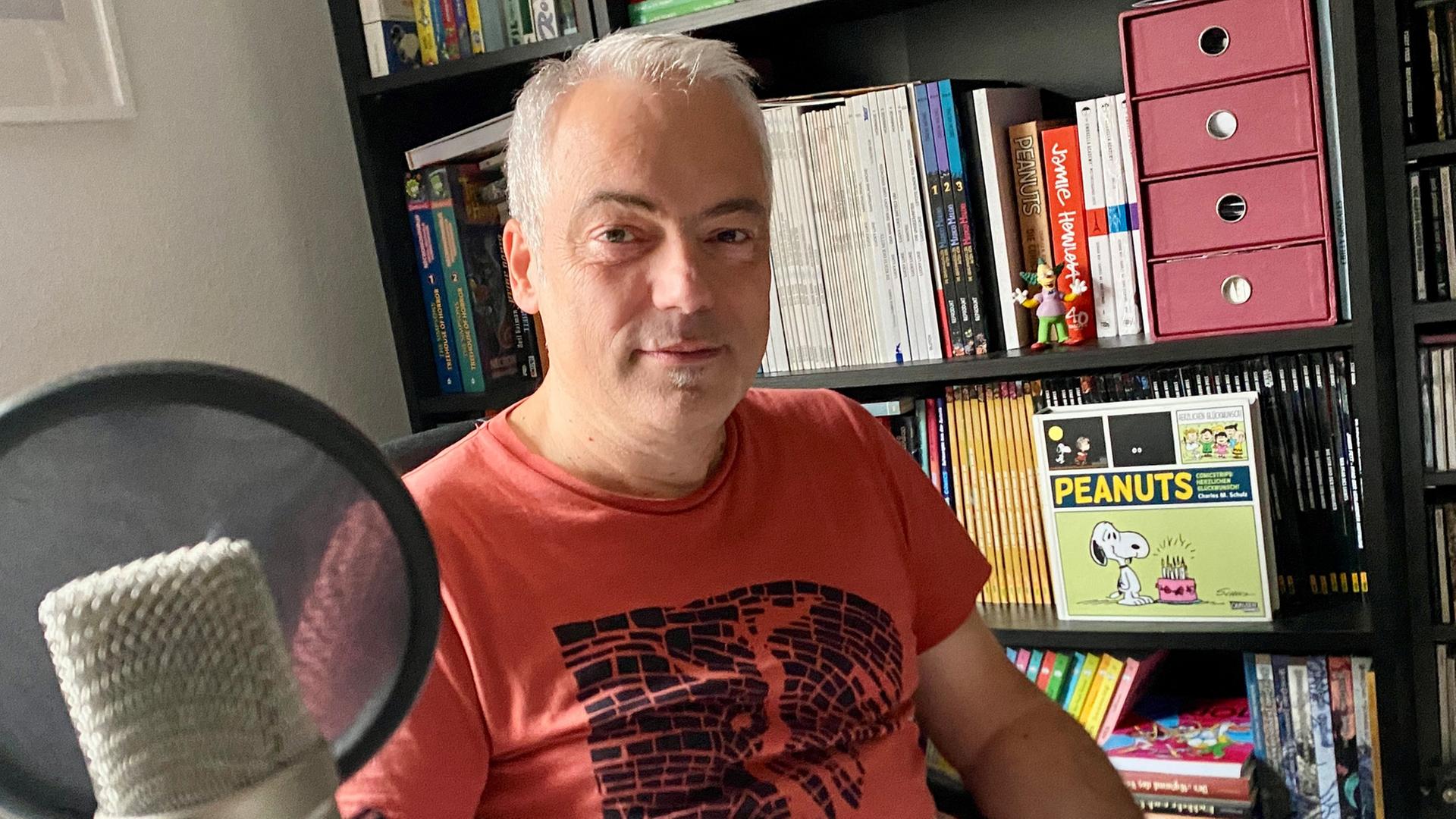 Ein Mann mit kurzen grauen Haaren sitzt vor einem Regal mit Peanuts-Hörbuch | NDR, Mathias Heller