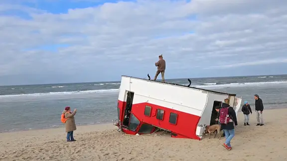 Das Schiffswrack ist für Strandwanderer nicht gesperrt. | Nachrichten ohne Unterbrechung An einem Strand auf der Nordseeinsel Sylt stehen Menschen um ein umgestürztes Lotsenboot.