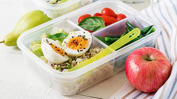 Eine Lunchbox mit Reis, gekochten Eiern, Tomaten und grünen Bohnen, daneben liegen ein Apfel und eine Birme.